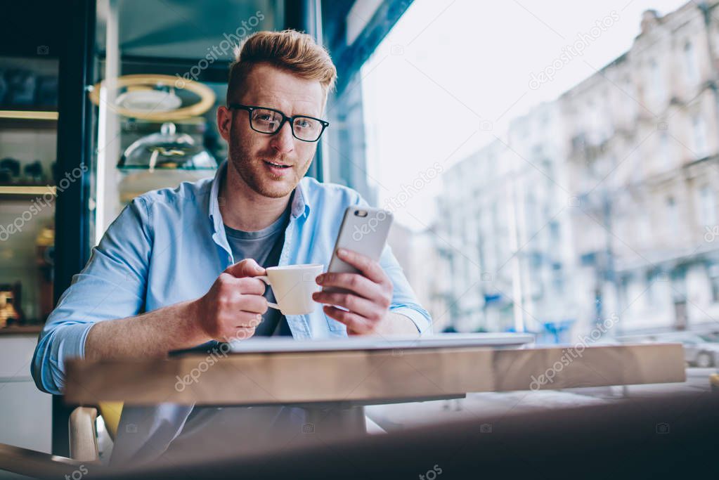 Portrait of smiling young man blogger enjoying tasty caffeine beverage while sharing media in social network on smartphone device using 4G internet connection.Positive hipster guy with cup of coffee