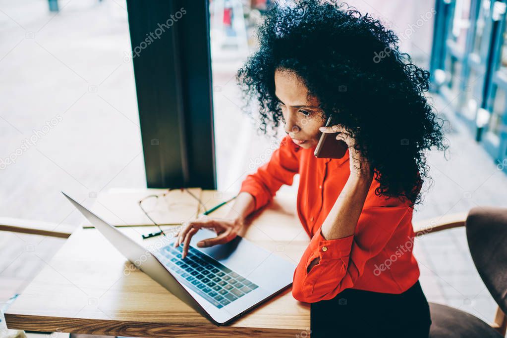Serious african american businesswoman talking with operator on smartphone while updating profile on modern laptop computer using wireless internet.Young woman calling on cellular sitting at netbook