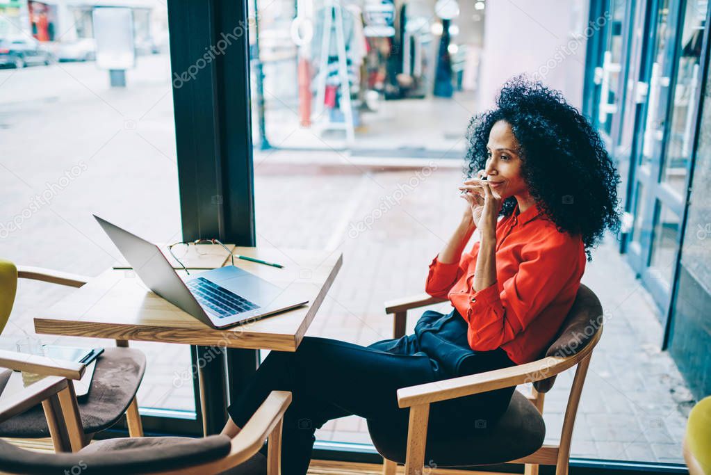 Positive dark skinned female graphic designer communicating during phone call on smartphone while sitting at table with modern laptop computer and working remotely in stylish coffee shop interior
