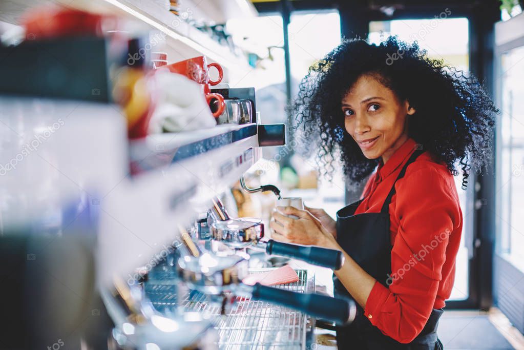 Portrait of successful african american young woman dressed in uniform working barista in own cafeteria and preparing tasty coffee beverage for client on modern machine developing own business