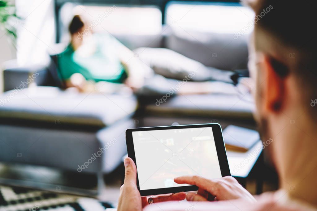 Cropped back view of young man pointing on blank screen of modern tablet and ignoring live communication with girlfriend which lying on couch.Selective focus on digital touch pad with copy space area