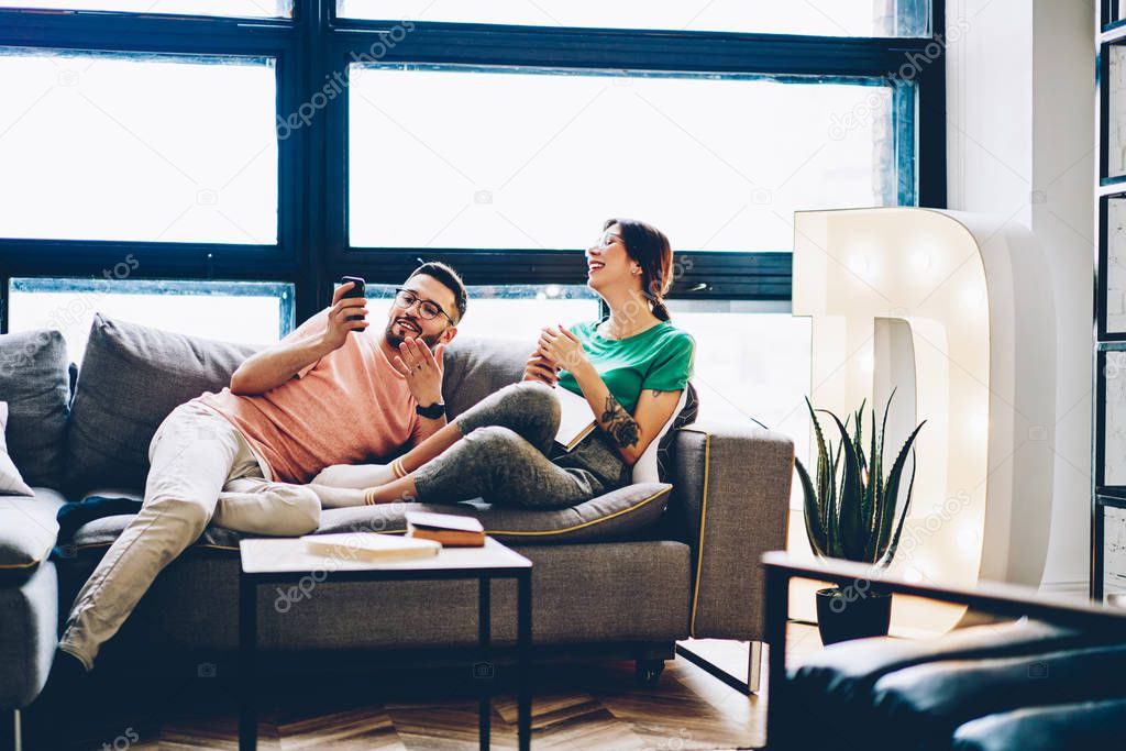 Young marriage resting on comfortable sofa and laughing during watching funny videos in blog on smartphone in stylish apartment.Positive couple in love discussing news in social networks on telephone