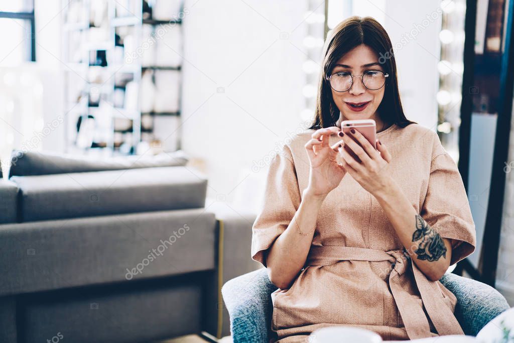 Positive casual dressed young woman in eyeglasses reading funny news on website using 4G internet on smartphone device.Cheerful hipster female blogger chatting with friends in social networks on phone