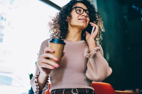 スマート フォンで 接続を使用して テイクアウトのコーヒーのカップを保持しているトレンディな眼鏡の女性を笑顔と携帯電話を介して通信 アプリケーション経由で友人と話して正千年内気な少女 — ストック写真