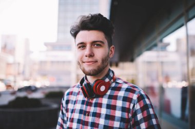 Handsoem genç adam rahat kıyafet ve bakarak sokak, hipster adam kamera ayakta açık havada eğlence keyfi kırmızı stereo kulaklık ile müzik dinleme için modern hassasiyet ile portresi
