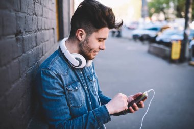 Download yeni şarkı ile kulaklıklar açık havada, serbest zaman sırasında caddenin üzerinde hücresel dinlenme üzerinde kısa mesaj gönderme olumlu hipster adam ayakta çalma listesi için telefon kullanan genç adam gülümseyen