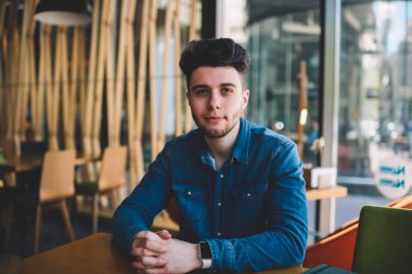 Yarım uzunluk portre yakışıklı kendine güvenen erkek Blogger Cafe iç, oturma rahat kot gömlek trendi saç modeli kamera kafeterya masada oturan bakarak adamla genç yenilikçi