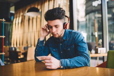 Kulaklık, hipster adam dinleme müzik playlist modern elektronik aksesuarlar ve holding kredi kartı üzerinden kafe iç masada oturan ziyaret kartındaki bilgileri kontrol ciddi genç adam