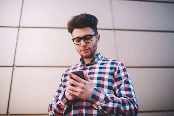 Positive hipster guy in trendy shirt using smartphone for chatting in social networks with friends ,handsome young man share multimedia content in blog using 4G connection and telephone apps