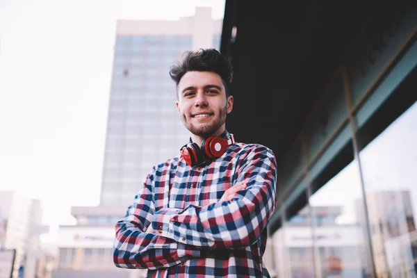 Half length portrait of confident hipster guy standing with crossed arms outdoors, handsome positive young man in stylish shirt and with modern red headphones looking at camera posting on urban setting
