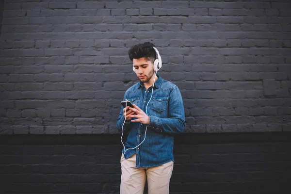 Pensive young man download song to smartphone playlist fond of music in stereo headphones,pensive hipster guy listening audio book in modern accessories connected to telephone standing on street