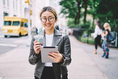 Yarım uzunluk portre çekici duygusal Asyalı kadın Blogger rekreasyon dijital tablet ve lezzetli içecek ile zevk. Kameraya bakarak ve Caddesi'nde yürürken gülen sevimli neşeli kadın