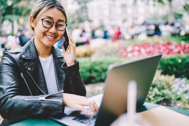 Telefonda konuşmak ve netbook üzerinde bilgi yazılacak gözlük başarılı Asyalı kadın yarım uzunluk portre. Neşeli kadın kişi teknoloji kullanırken kameraya bakarak kırpılmış görüntü
