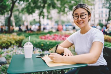 Yarım uzunluk portre çekici gülümseyen akıllı kadın yazarın fikirleri yayın hakkında daha iyi görme durulması için gözlük. Kameraya Park'ta otururken müreffeh hippi kız