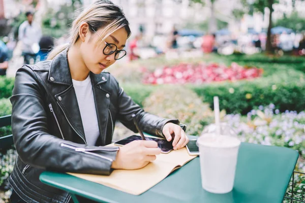 Young gorgeous creative female blogger in glasses for better vision recording ideas while spending free time at street.Serious hipster girl planning schedule writing down information to netbook