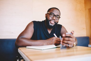 Cep telefonu tutan ve Not Defteri'nde yazma gözlük neşeli Afro-Amerikan hipster adam portresi, Genç karanlık gülümseyen sakallı adam notlar ve ağ kameraya bakan derili