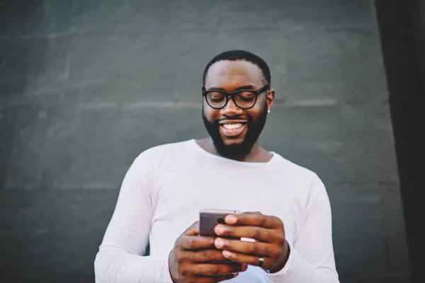 Cheerful african american hipster guy reading information from cellular browsed web page standing outdoors, smiling dark skinned male happy about get new income message using roaming internet