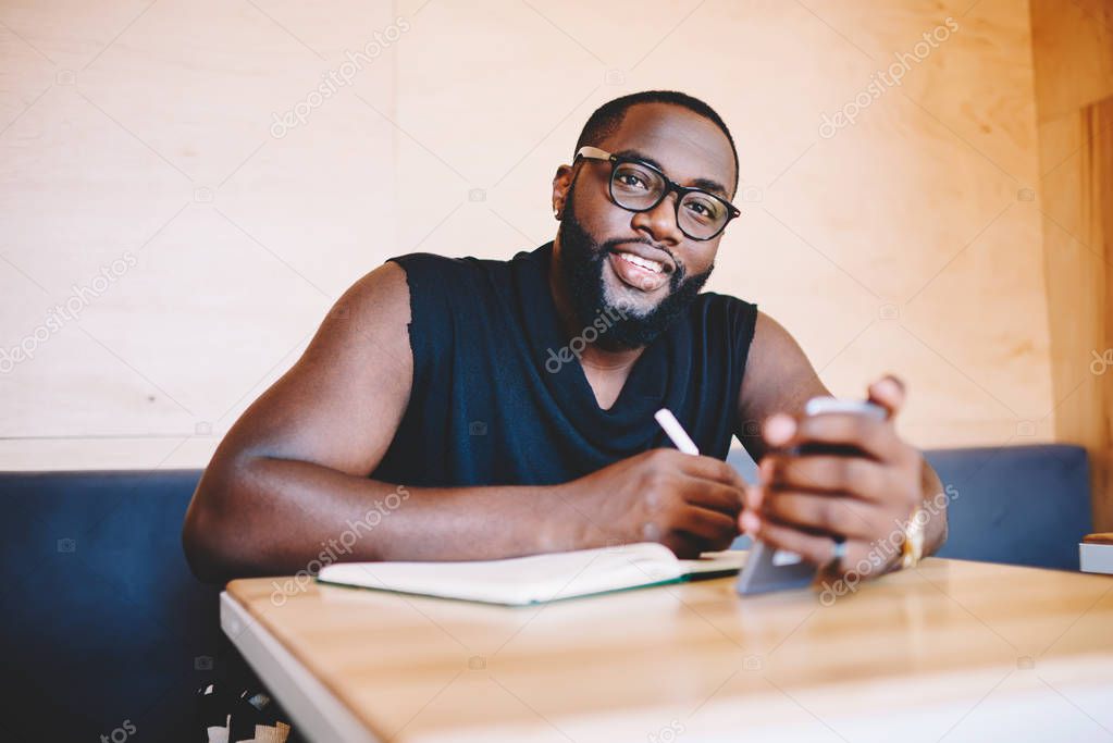Portrait of cheerful african american hipster guy in spectacles holding mobile phone and writing in notepad, smiling young dark skinned bearded man looking at camera making notes and networking