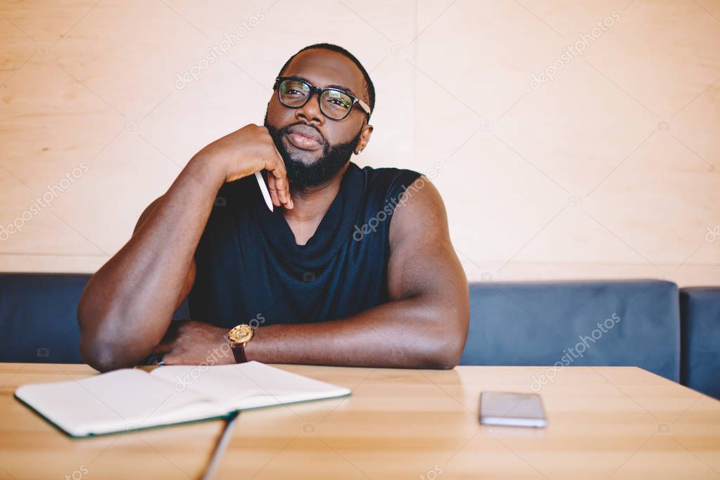 Pensive african american bearded guy in eyewear looking away dreaming about something while sitting at cafe interior with notepad, serious darks skinned man thinking about ideas for project