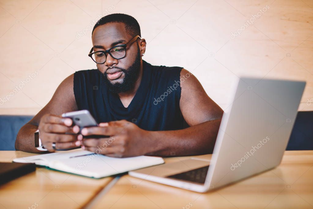 Serious african american male freelancer reading information from web page on telephone,young dark skinned businessman in eyewear checking banking balance via smartphone app sitting with laptop