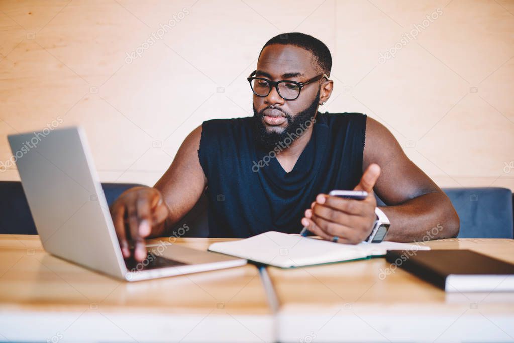 Serious dark skinned male freelancer browsing app on laptop computer during remote job, skilled african american student using modern technology and wifi while learning online in cafe interior