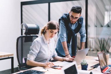 Masaüstünde oturan ve akıllı telefon kullanan konsantre kadın yönetici, beyaz erkek çalışanın yanında durup bilgi bekliyor. İki iş arkadaşı iş çözümlerini analiz etme ve arama