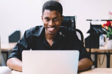 Neşeli erkek yazılım uzmanı portre kamera bakarak ve birlikte çalışma ofis masada ön dizüstü bilgisayar otururken gülüyor, mutlu ve başarılı Afrikalı Amerikalı adam masaüstü çalışan