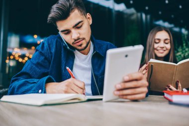 Yoğunlaşmış kafkas erkek, kadın arkadaşı kitap okurken, yoğun genç adam araştırma için modern teknolojiyi kullanırken, telefon görüşmesi ve bilgi notları yapma rezervasyon onaylayan