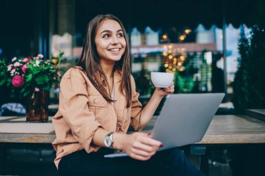 Neşeli esmer kadın hipster zevk aroma kahve molası fincan tutan ve uzakta gülümseyerek bakarak, yetenekli başarılı kadın freelancer kafede dizüstü bilgisayar da uzak iş yapıyor