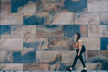 Reklam veya metin mesajları için kopya alanı ile sokakta kentsel ayar duvar geçen sırt çantası ile kadın genç öğrenci, tanıtım alanı yakınında yürüyüş rahat trendy kıyafeti genç hipster kız