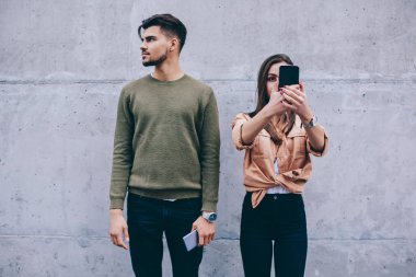Genç yakışıklı adam gerçek iletişim görmezden gelirken kız arkadaşı akıllı telefon üzerinden online sohbet tercih ederken, hipsters yüzünün önünde hücresel tutan kadın duvarda duran