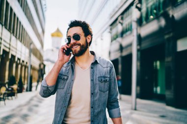 Denim mutlu sakallı hipster adam boş zaman zevk alışveriş merkezinde yürüyüş akıllı telefon konuşma sırasında gülerek giymek. Güneş gözlüğü pozitif genç adam cep telefonu açık havada gezinen arama