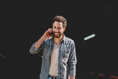 Yakışıklı sakallı genç adamın, otoparkta dart duran akıllı telefon cihazını ararken kameraya gülümseyen portresi. Hücresel konuşurken rahat giyim giymiş iyi görünümlü hipster adam