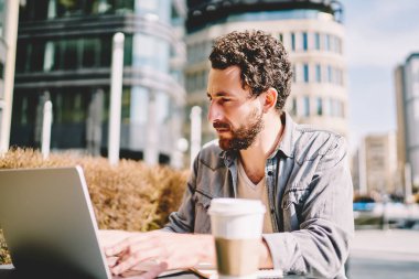 Pensive sakallı serbest dizüstü bilgisayarda oturan ve kafe açık havada ücretsiz 4g internet kullanarak internet web sitesinde grafik gelişimi üzerinde çalışıyor. Netbook'ta haber okuyan konsantre genç adam