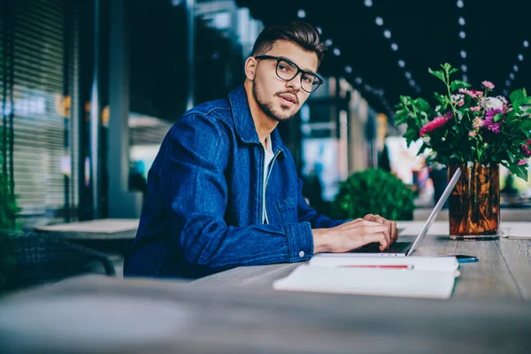 stock image Portrait of serious caucasian male in spectacles working remotely on laptop computer, pensive hipster guy in spectacles for vision correction keyboarding publication for blog looking at camera