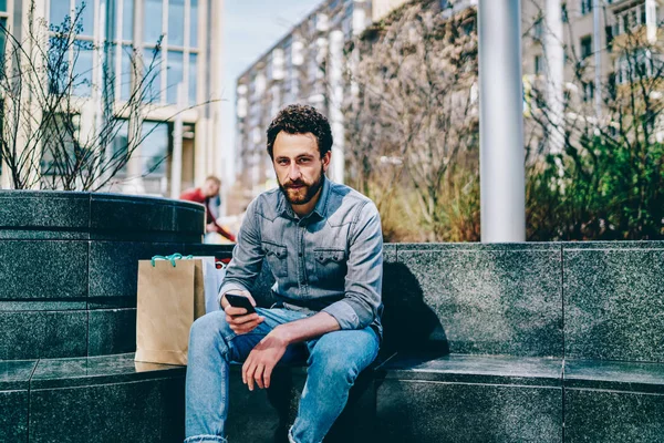 Portrait of bearded hipster guy dressed in denim wear holding smartphone device and updating profile in social networks while resting with paper bags after good shopping in center of modern city