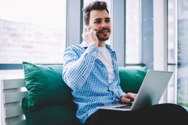 Rahat giyim beyaz adam bacaklarüzerinde dizüstü bilgisayar ile oturan ve uzaktan çalışmak için 4g kablosuz bağlantı kullanarak mobil uygulama üzerinden akıllı telefon konuşma sırasında bilgi tartışıyor