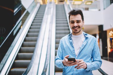 Elinde modern akıllı telefon gadget ile elektrikli merdivenlerde ayakta ve boş zamanlarında iyi hissederken kamera bakarak şık gömlek mutlu gülümseyen hipster adam yarım uzunlukta portre