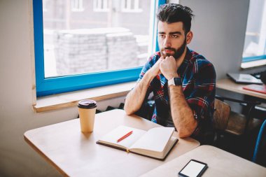 Ciddi sakallı hipster öğrenci dizüstü bilgisayar ile masaüstünde otururken düz görünümlü ve akademide ev ödevi düşünüyorum. Gündelik giyim genç adam eğitim ve üniversitede seminer için hazırlanıyor