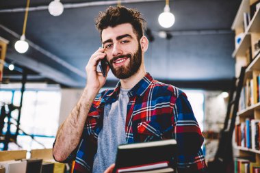 Üniversite kütüphanesinde duran kameraya gülümserken modern akıllı telefonda konuşan yakışıklı neşeli genç adamın portresi. Pozitif hipster öğrenci edebiyat kitapları tutan telefonda arama