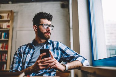 Akıllı telefon cihazındaki sosyal ağlardaki profili güncellerken gözlüklü hipster öğrencisinin pencereden dışarı bakmasından. Düşünceli sakallı genç adam cep telefonu online sohbet