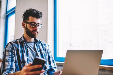 Kablosuz internet kullanarak modern dizüstü bilgisayarda web semineri izlerken pensive sakallı geliştirici elinde akıllı telefon tutarak. Genç adam üniversitede oturan netbook üzerinde uygulama yükleme