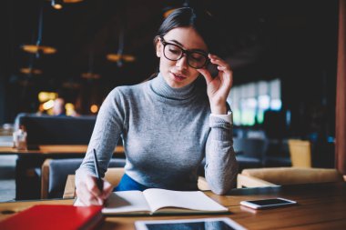 Zeki kız öğrenci 20 yaşında üniversite kütüphanesinde oturan ve ders kitabı ile sınava hazırlanıyor, genç akıllı hipster kız not defterinde başlangıç projesi hakkında izlenim incelemesi yazma