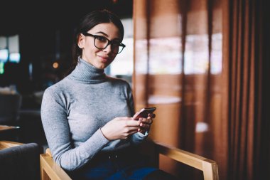 Cep telefonu ile yeni bir yayın oluştururken gözlük neşeli gülümseyen blogger Portresi, ortak çalışma alanında dinlenen modern cep telefonu teknolojisi tutan mutlu hipster kız