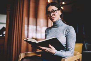 Neşeli kız öğrenci 20 yaşında esmer saçlı kapalı oturan ve dizüstü tutan, şık kıyafeti genç pozitif hipster kız rahat kafeteryada edebiyat ile serbest zaman zevk