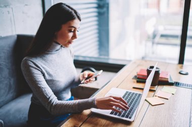 Düşünceli iş kadını 20s kahve dükkanında oturan web mağazasında alışveriş için dizüstü bilgisayar kullanırken modern akıllı telefon uygulaması üzerinden ödeme yaparken, teknoloji ve iletişim kavramı