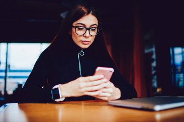 Modern dizüstü bilgisayarda eğitim mola wifi bölgesine bağlı akıllı telefon üzerinden bildirim kontrol Pensive hipster kız, görme düzeltme arkadaşları ile sohbet için gözlük güzel kadın