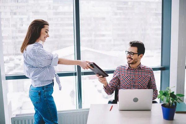 Positive cheerful man dressed in casual wear sitting at company desktop with modern laptop computer and taking textbook from happy successful female partner standing near in office environment