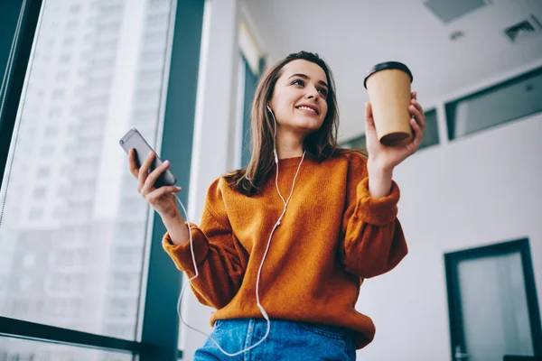Positive hipster girl smiling during listening motivation audio book via electronic headphones and online application for education holding coffee to go indoors, happy woman enjoying time for music