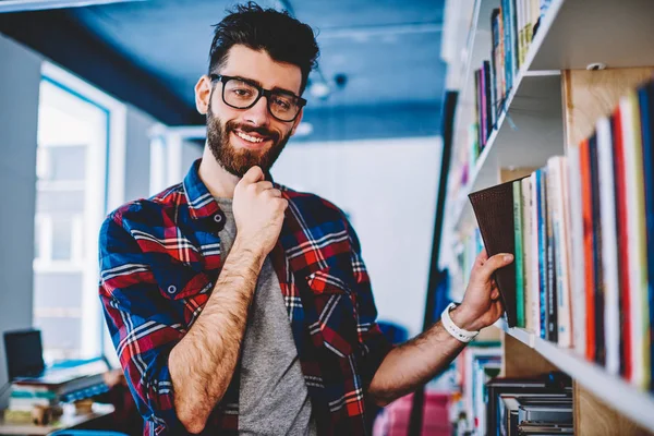 在大学的书架上挑选文学书籍时 戴着眼镜的留胡子的时髦学生在镜头前微笑 正心的年轻人在休闲衬衫站在图书馆 — 图库照片